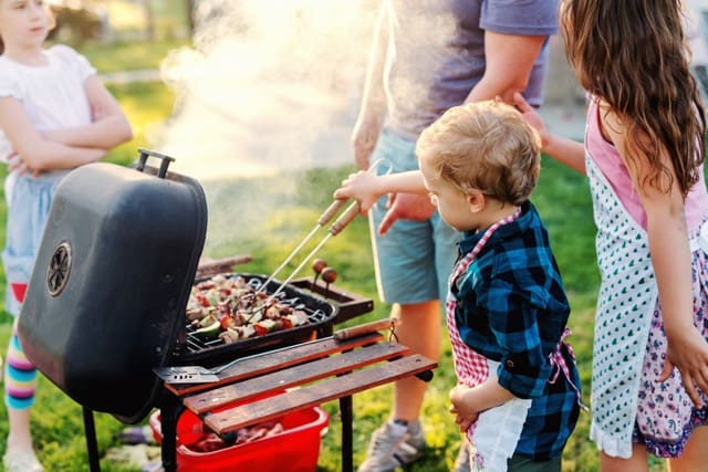 Little chef with apron grilling meat and vegetables on sticks Next to him his cousins Family gathering concept
