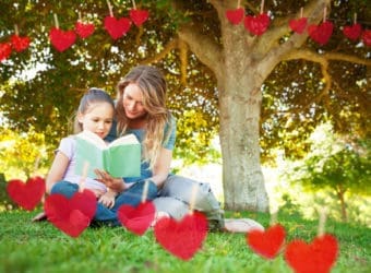 Mother and daughter reading a book at park against hearts hanging on a line